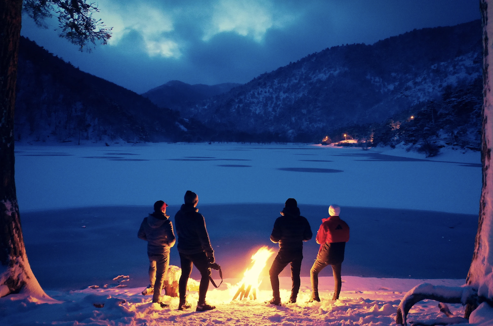 Four people at a campfire looking at the lake in front of mountains at night.